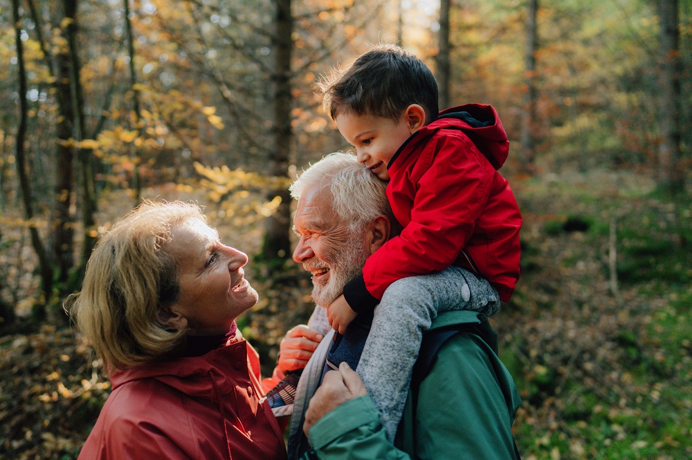 Toddler sitting on his grandad's shoulder smiling at his grandma, outdoors in woodland