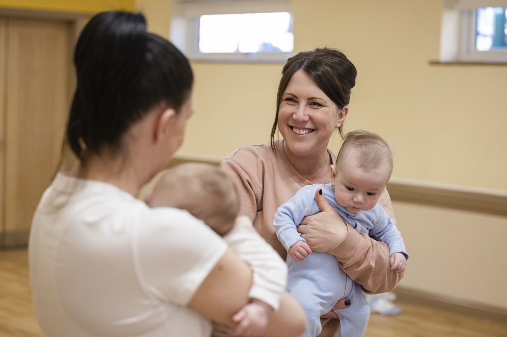 Two mums holding babies chatting and smiling in a community hall