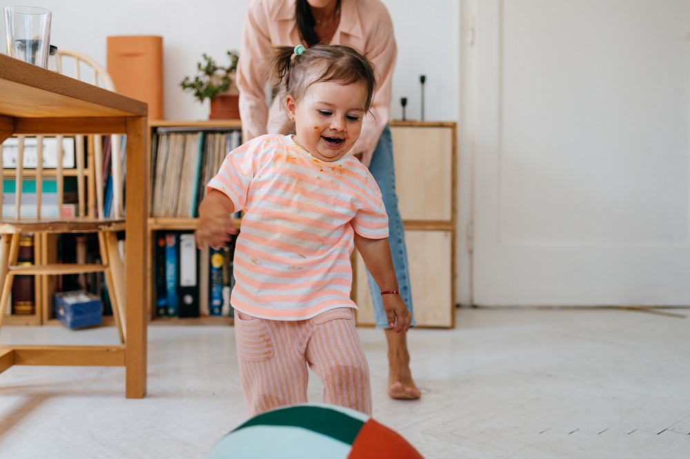 Happy toddler girl running towards a ball in a living room, supported by her mum