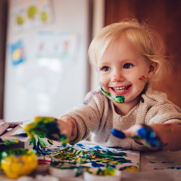 Image of a smiling child with colourful paint on their hands and face.