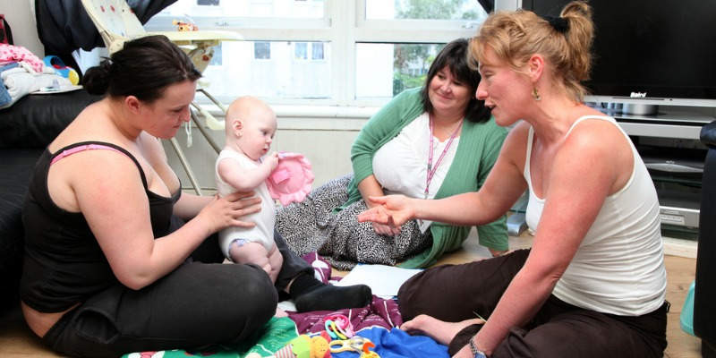 Image of three women sitting in a circle on the floor, looking at a baby and smiling.