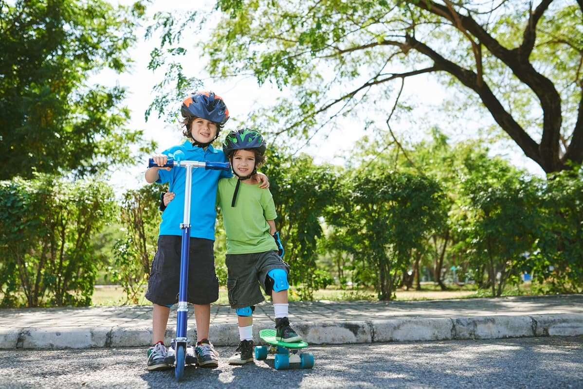 Two boys, one with a scooter, one with a skateboard, both wearing helmets