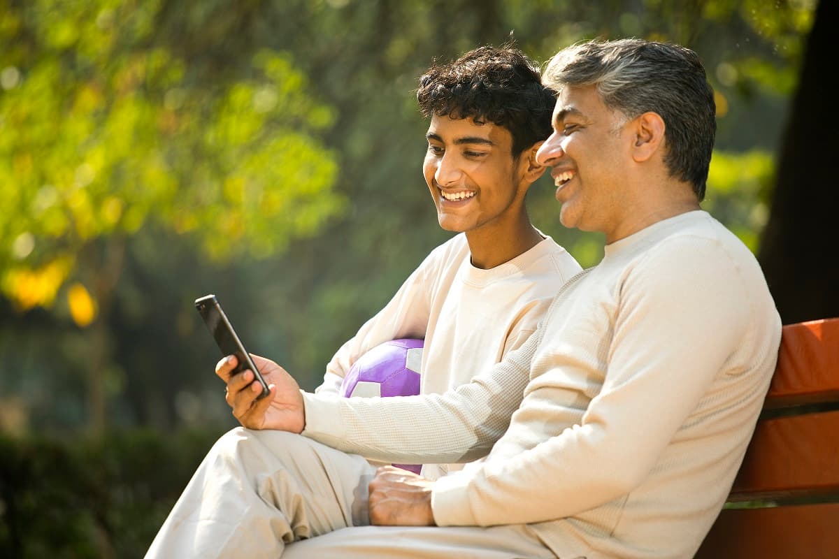 Dad and teen boy sitting on a bench outside looking at a mobile phone and smiling