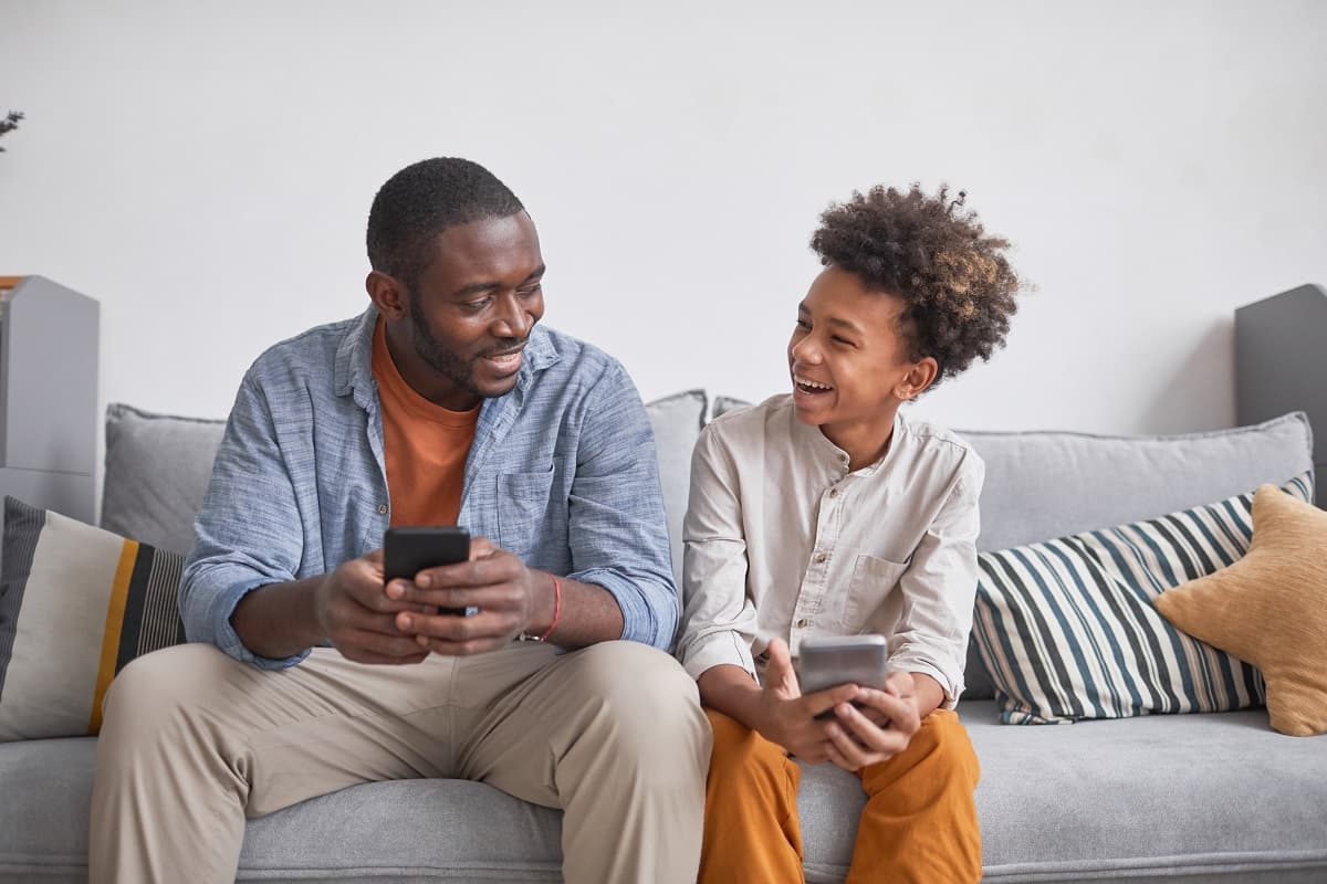 Dad and teen boy sitting on sofa holding phones, looking at each other and laughing