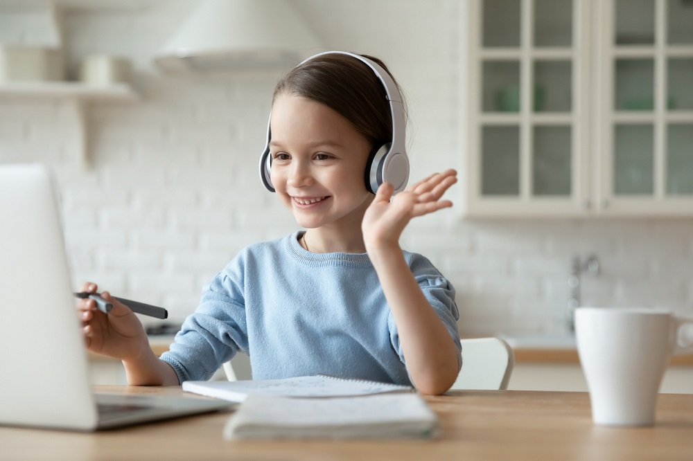 Little girl wearing headphones learning online and waving to the laptop screen