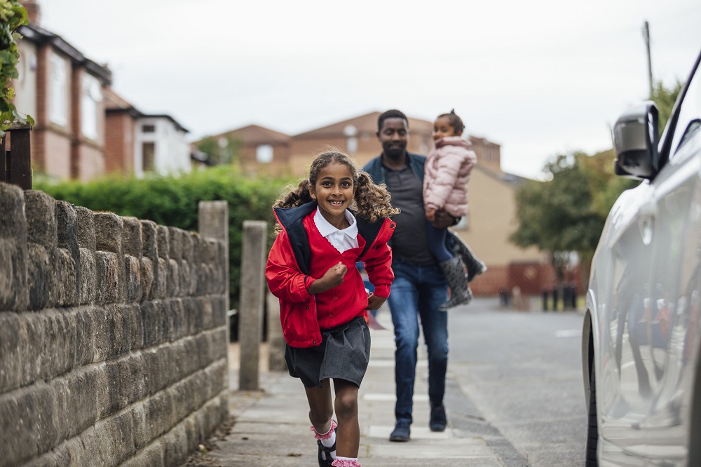 Young girl carrying a backpack running into school, her dad carrying a toddler walking behind