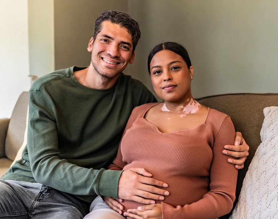 Pregnant couple sitting on a sofa, the dad with his hand on the mum's stomach.