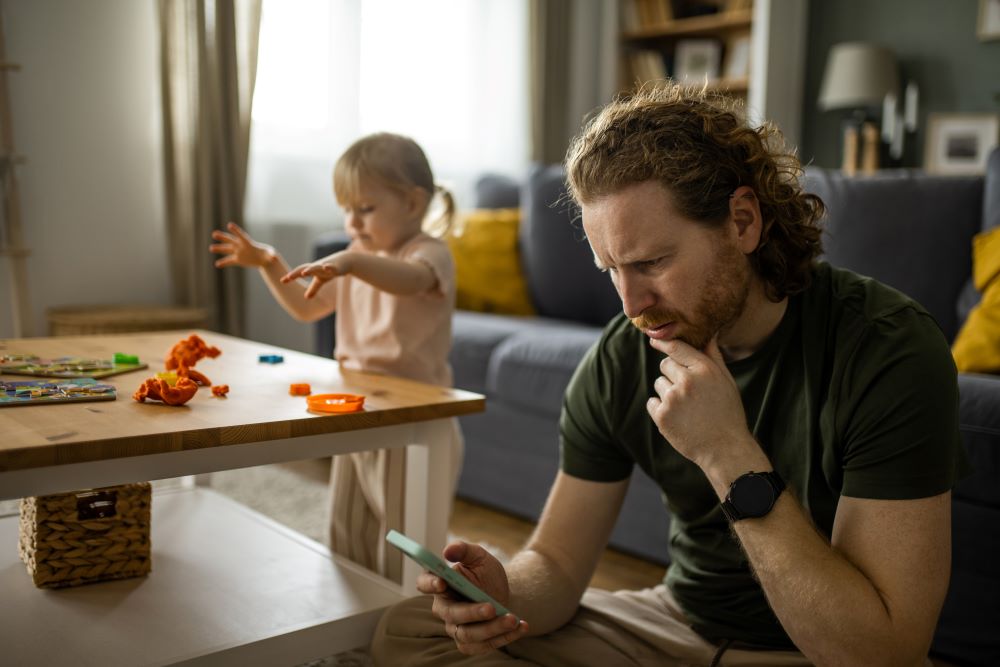 Anxious dad looking at his phone will his child plays with blocks in the background.