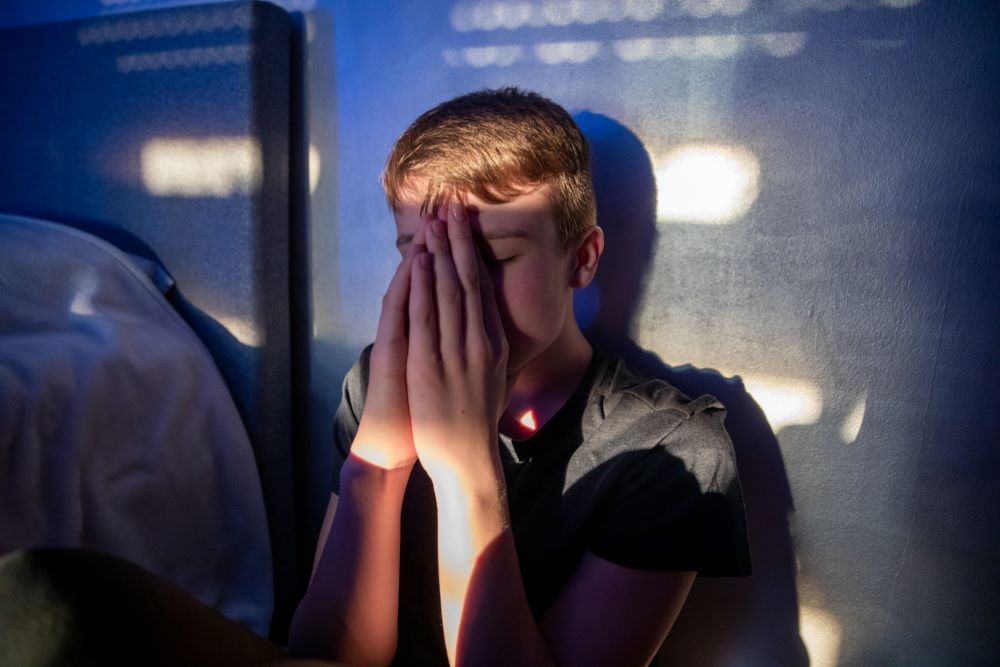Anxious teenage boy in a dark room covering his face with his hands.