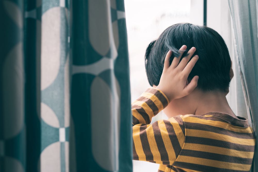 Little boy standing by a window with his hands over his ears.