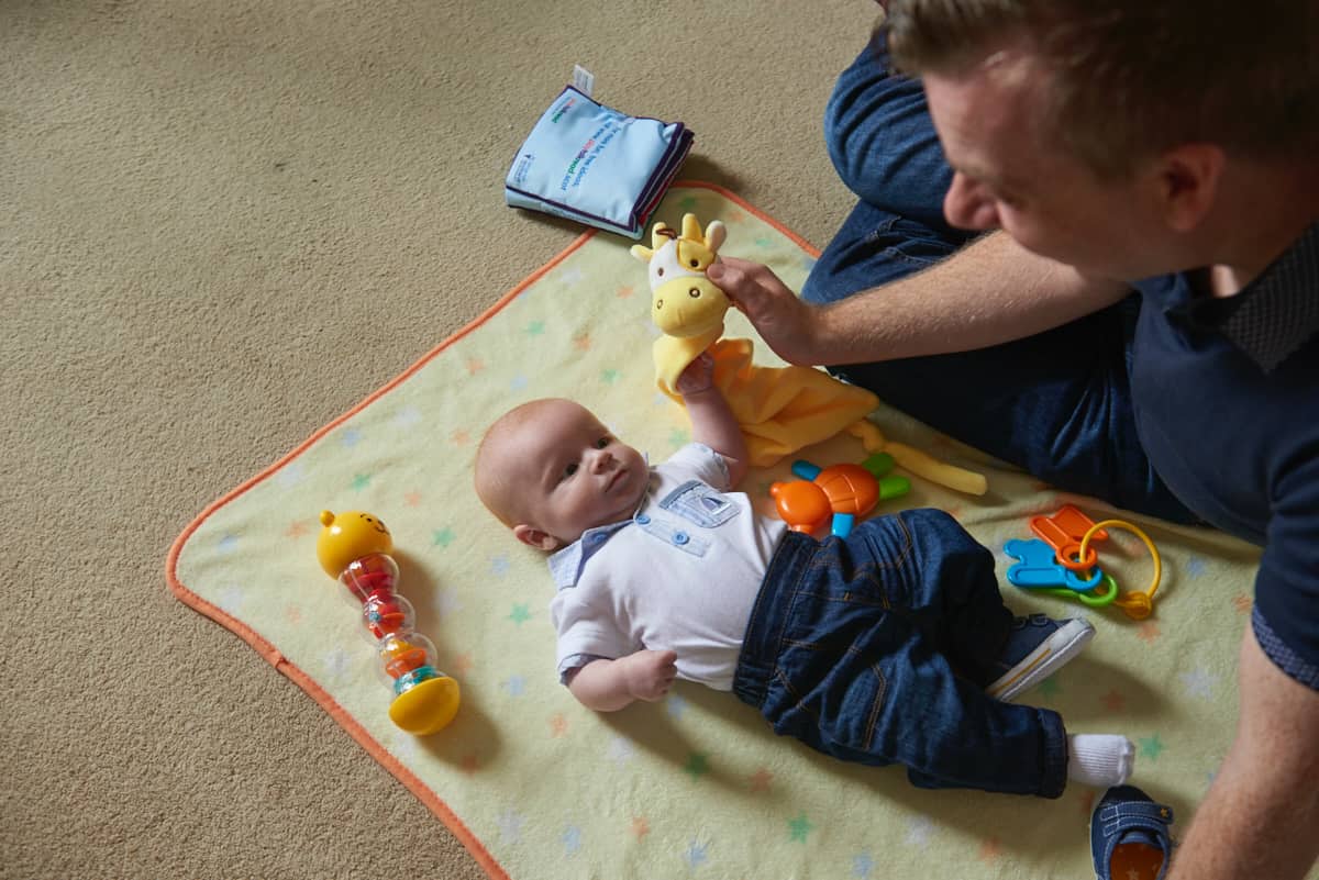 Image of a baby lying on a mat looking up at their dad, who is holding toys for the baby to play with.