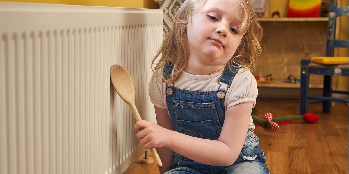 Image of a child sitting on the floor next to a radiator, holding a wooden spoon.