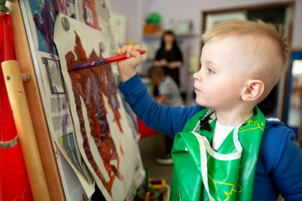 Image of a child standing in a playroom, painting on a piece of paper.