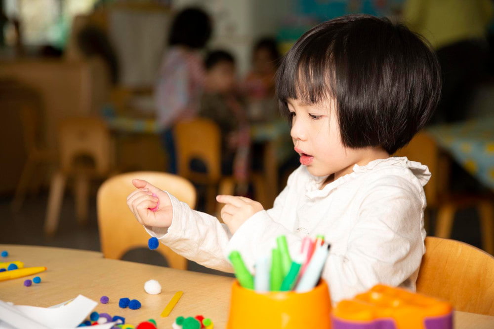 Image of a child sitting at a table in a playroom, doing arts and crafts using small pom poms.