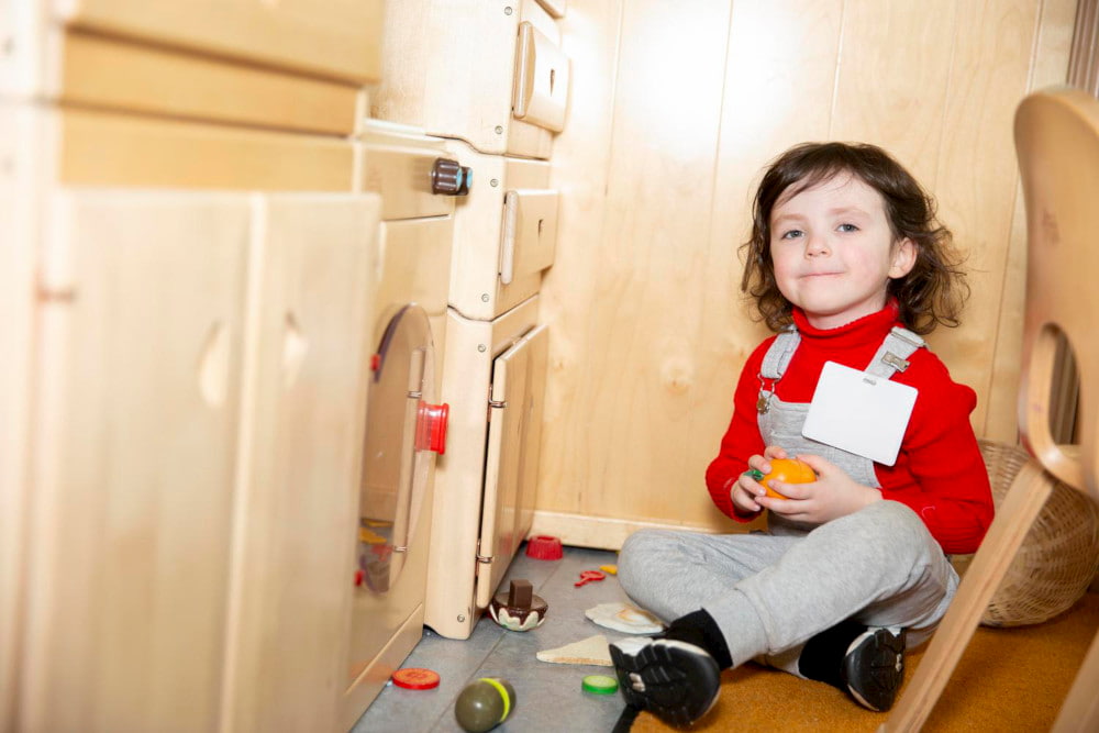Image of a child sitting on the floor of a playroom, playing with miniature kitchen toys.