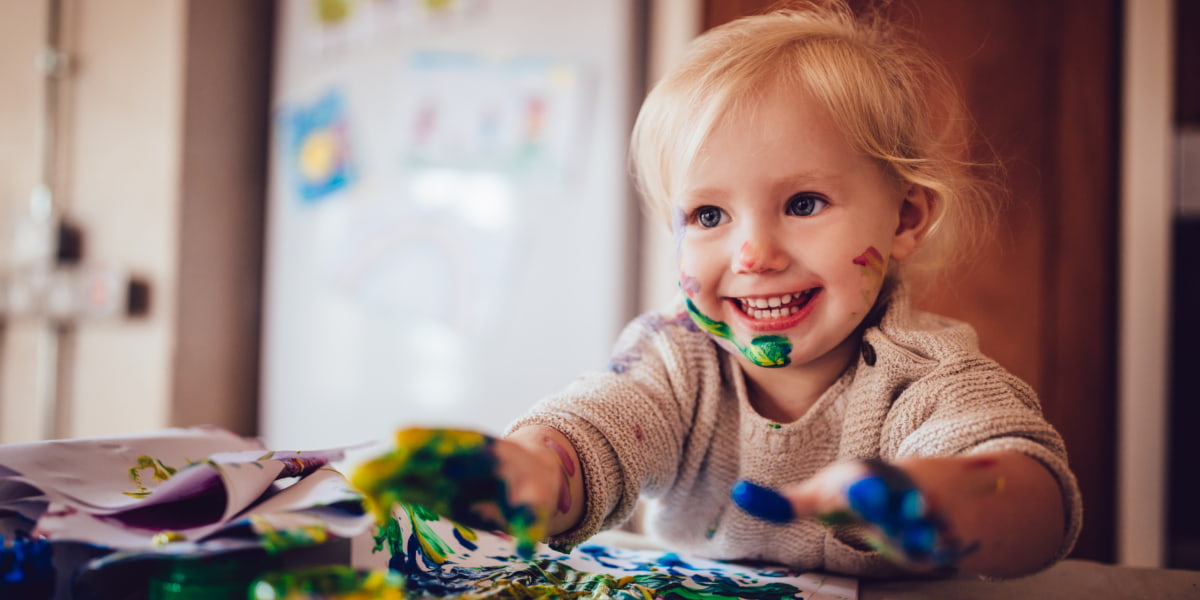 Image of a smiling child with colourful paint on their hands and face.