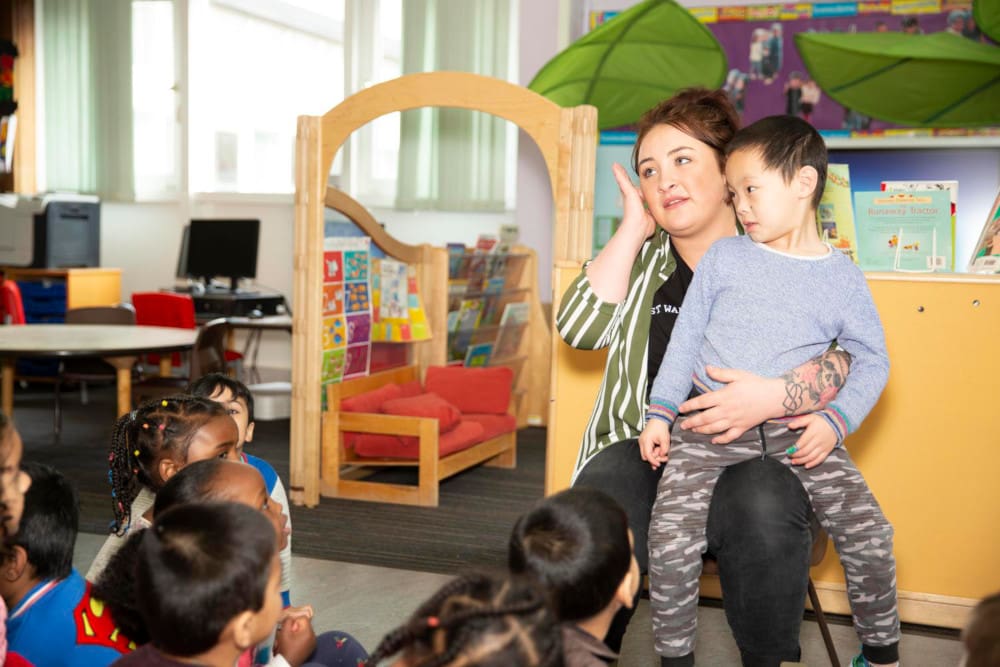 Image of children sitting on the floor of a playroom looking at a teacher holding a boy.