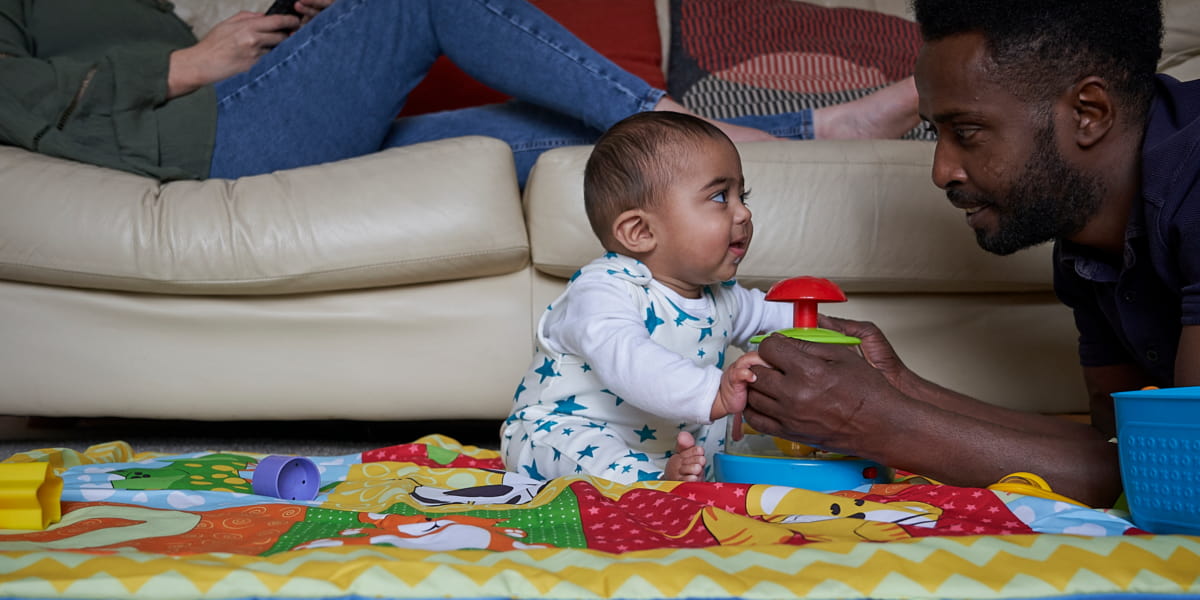 Image of a dad and baby smiling at each other while they play with toys on a mat on the floor.