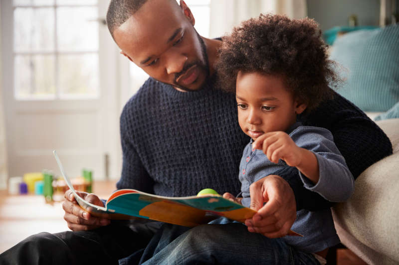 Image of a dad and child sitting together on a sofa reading a book.