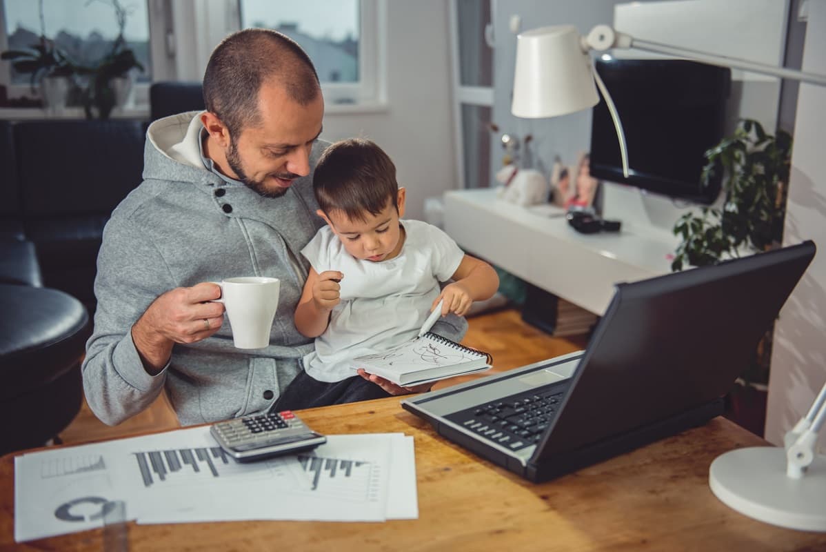 Dad with toddler on his knee, looking at forms and a laptop