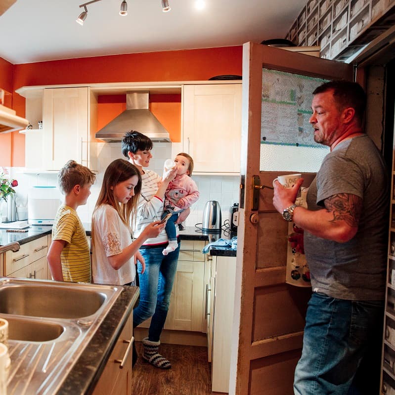 Family in the kitchen, mum holding a baby, dad coming through the kitchen door, young boy and girl looking at a phone