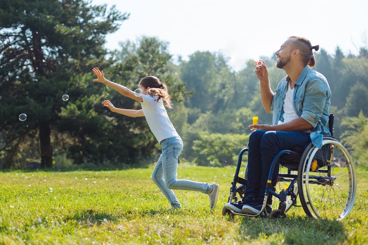 Dad blowing bubbles for his daughter in the park; dad is in a wheelchair