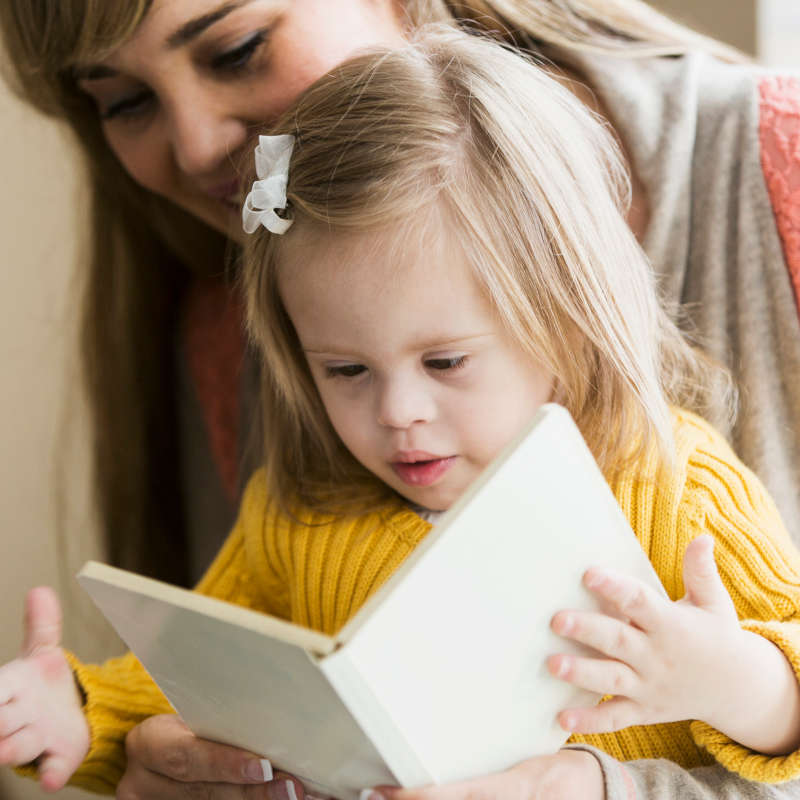 Image of a child sitting on their smiling mum's lap and looking at a book together.