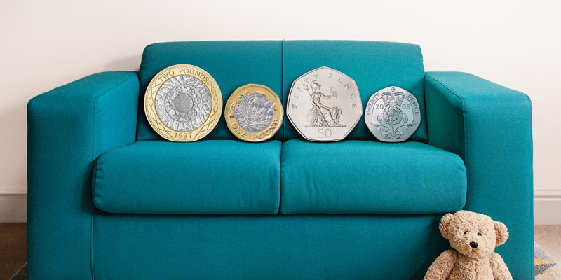 Image of a sofa with four giant coins sitting on it and a teddy bear sitting in front of the sofa.