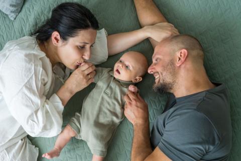 Mum and dad lying on a bed with their newborn baby, looking at the baby lovingly.