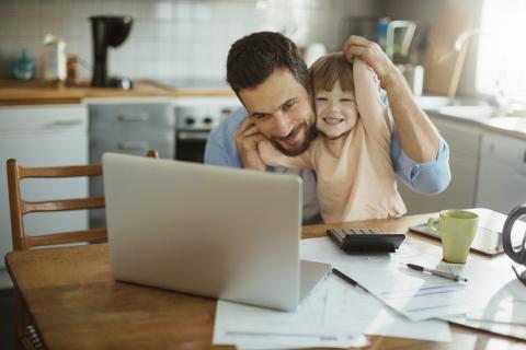 Toddler sitting on dad's knee, looking at a laptop and calculator, with papers around them