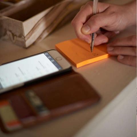 Image of a hand writing on a post-it note with a phone and a wallet next to it on a table.