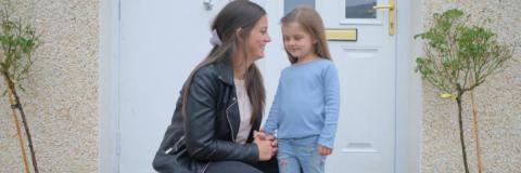 Image of a mum crouching down next to her child and smiling at them in front of their front door.