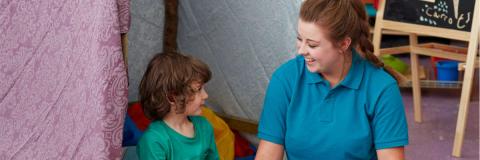 Image of a staff member and a child sitting together on the carpet of a playroom, smiling at each other.