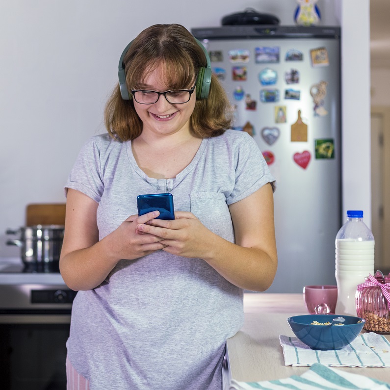 Teen girl in the kitchen wearing headphones and looking at her phone smiling