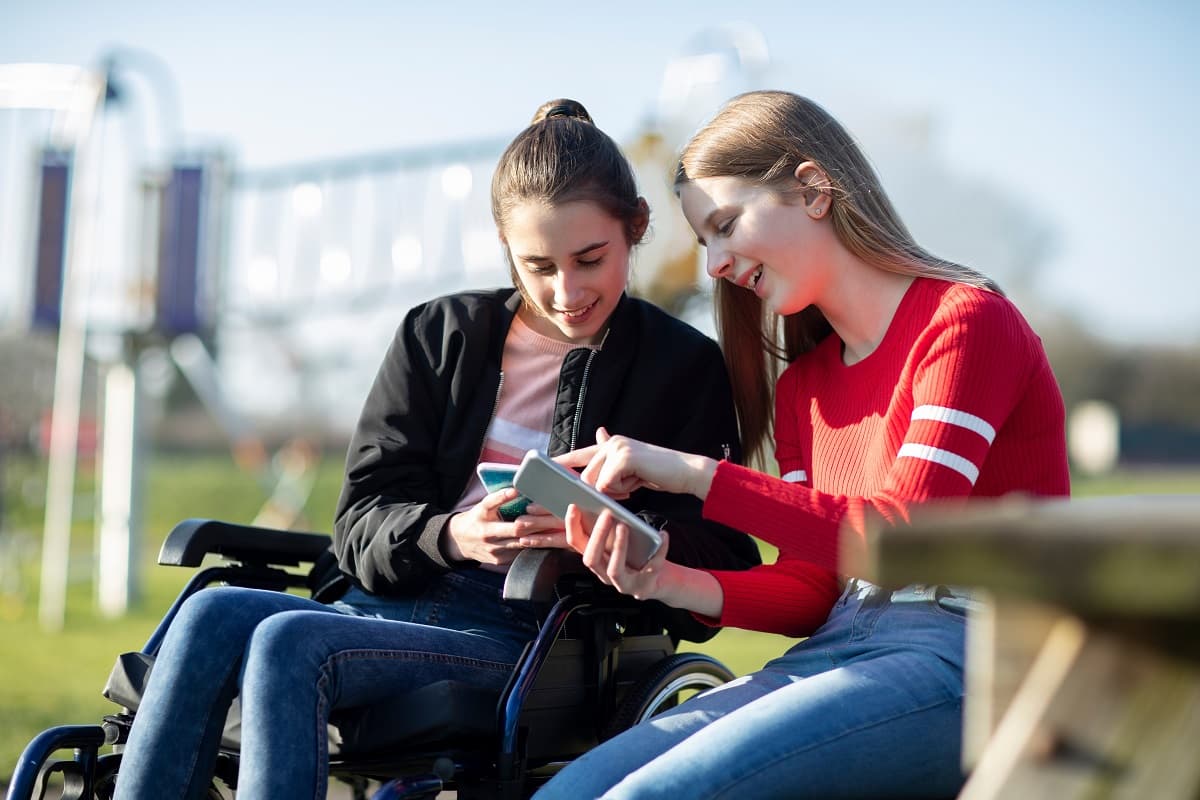Two teen girls outdoors, looking at their phones. One is in a wheelchair.