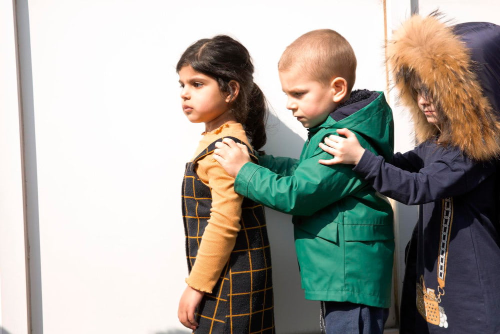 Image of three children standing in a line with their hands on the shoulders of the child in front of them.