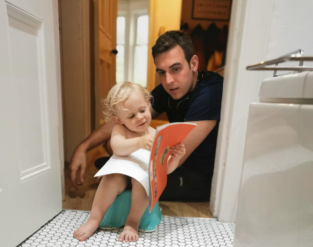 Image of a toddler sitting on a child's toilet reading a book, with their dad sitting near them.