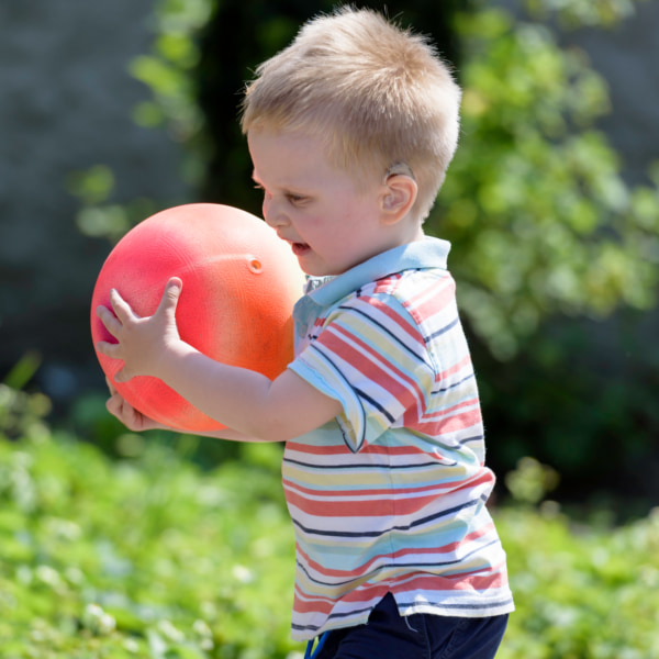 Image of a toddler in a garden, holding a red ball and smiling.