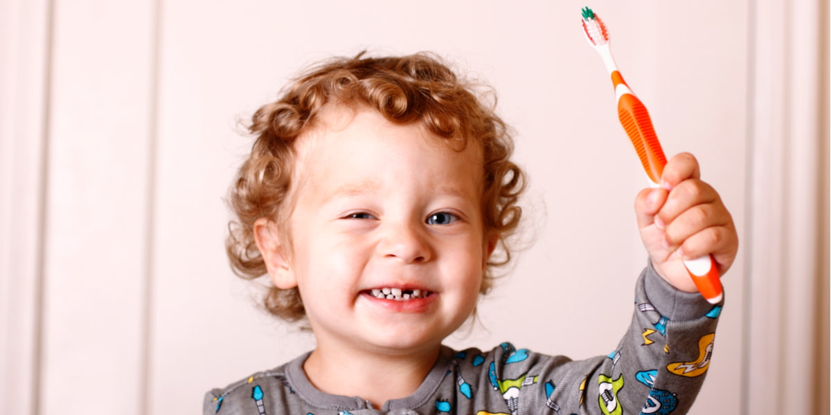 Image of a toddler smiling at the camera and holding a toothbrush up in the air.