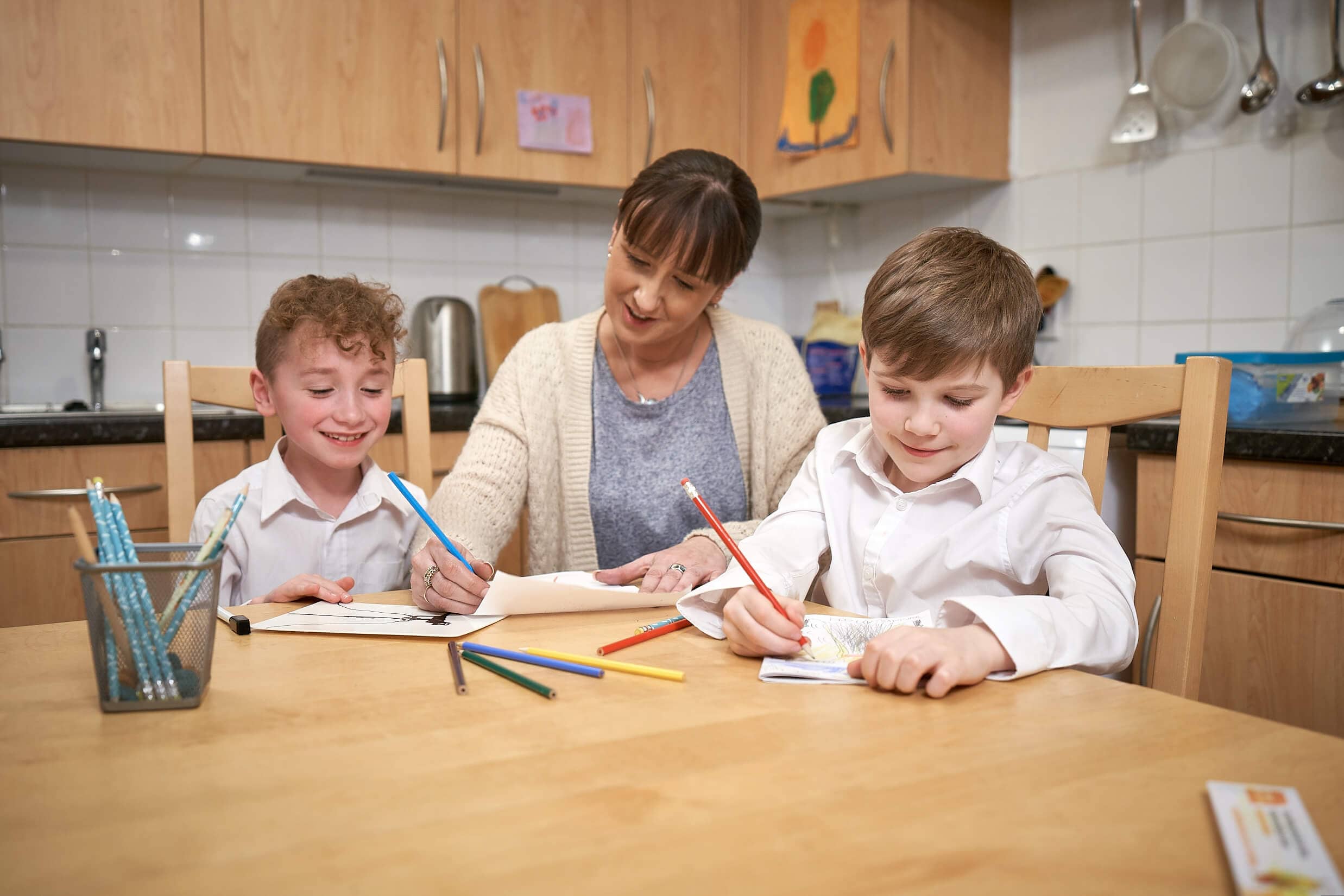 Image of two children sitting at a kitchen table with their mum, smiling and drawing with colouring pencils.