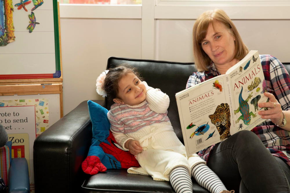 Image of a woman reading to a child, sitting next to each other on a sofa in a playroom.